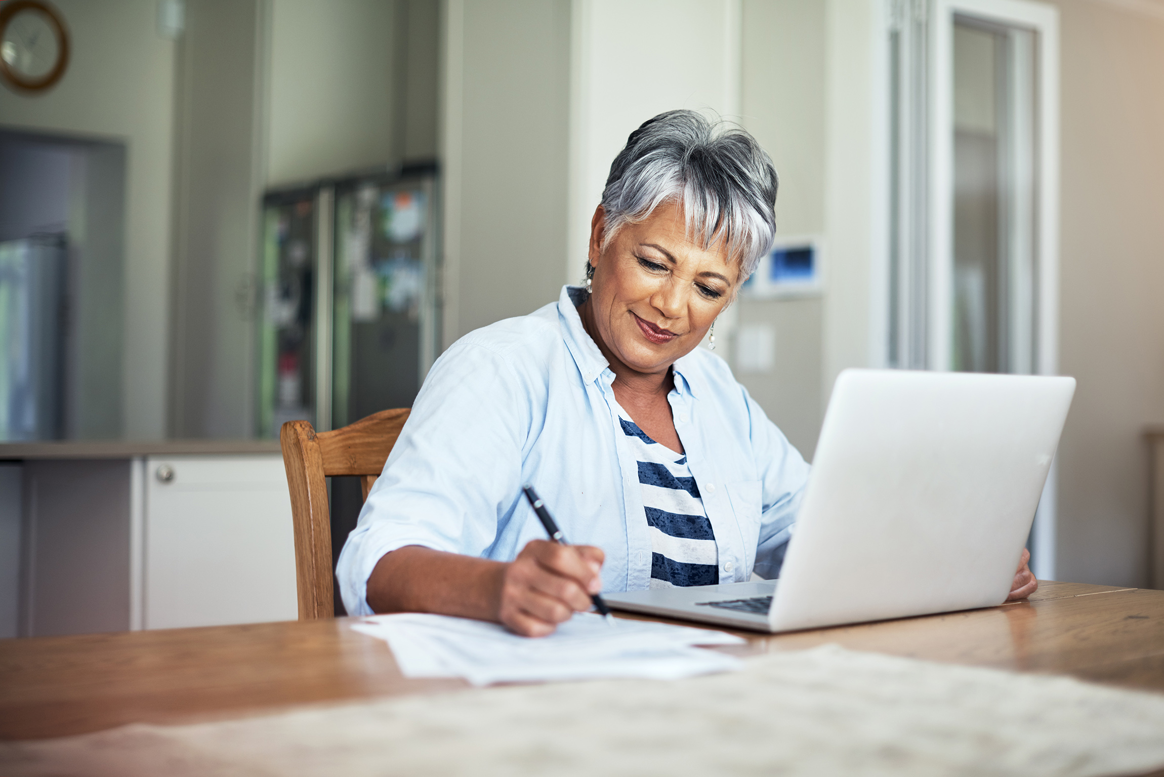 An older woman uses a laptop while taking notes with pen and paper.