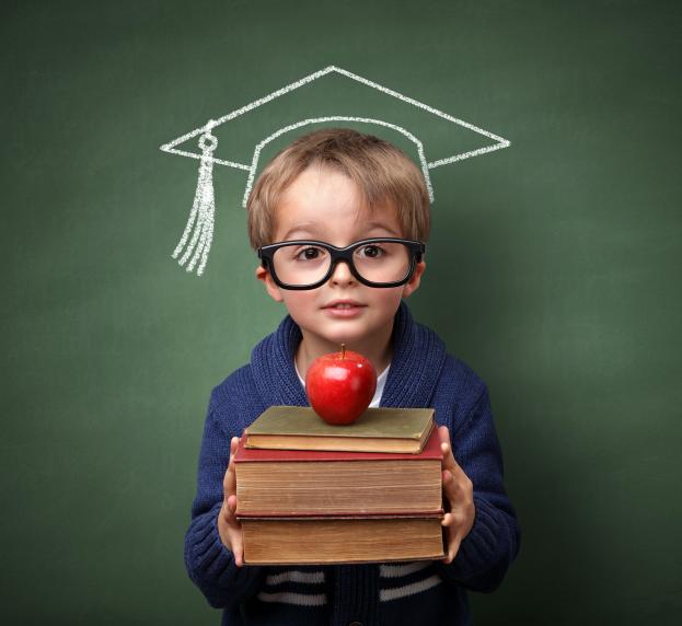 A young boy poses with books, an apple, and a chalk graduation cap.